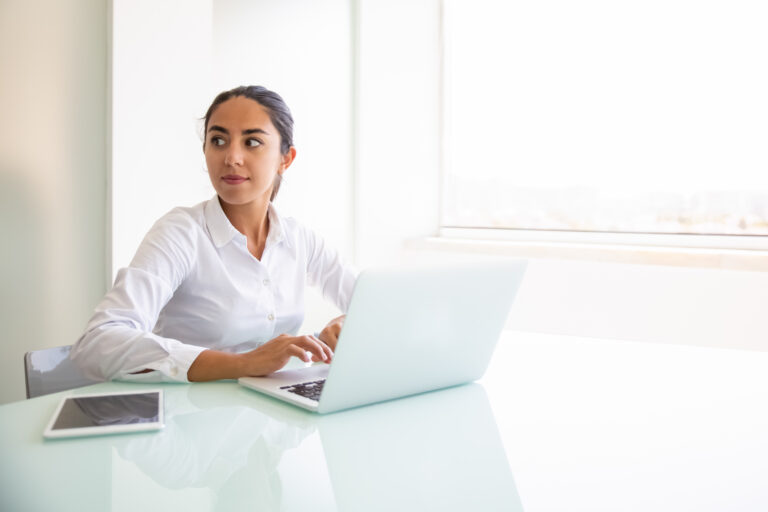 Confident female consultant working on computer. Young Latin businesswoman sitting at workplace, using laptop, looking away. Professional at workplace concept