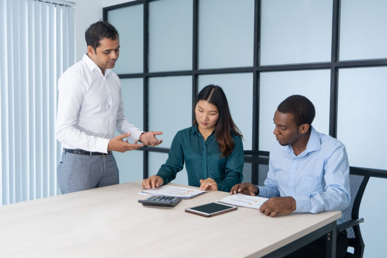 Experienced employee instructing newcomers. Asian and Afro American office workers studying documents at conference table, Indian supervisor explaining details. International team and training concept