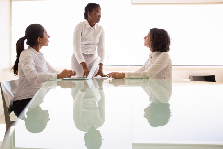 Female business team discussing project in conference room. Businesswomen meeting at table, using laptop, talking, sitting, standing. Corporate meeting concept